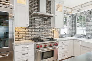 Kitchen with light stone counters, white cabinetry, glass insert cabinets, wall chimney range hood, and designer stove