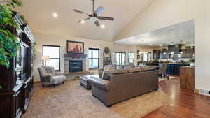 Living room featuring high vaulted ceiling, visible vents, a stone fireplace, ceiling fan with notable chandelier, and dark wood-style floors