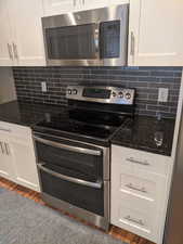 Kitchen featuring decorative backsplash, white cabinets, stainless steel appliances, and dark stone counters.