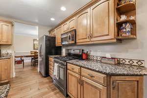 Kitchen with open shelves, light wood-style flooring, dark stone countertops, stainless steel appliances, and ornamental molding