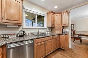 Kitchen featuring light wood finished floors, ornamental molding, dark stone counters, a sink, and stainless steel dishwasher