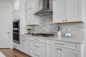 Kitchen with white cabinetry, tasteful backsplash, wood finished floors, stainless steel appliances, and wall chimney range hood