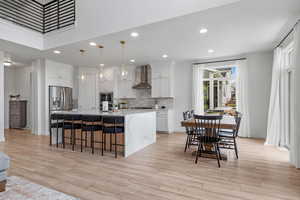 Kitchen with white cabinetry, wall chimney exhaust hood, a kitchen bar, stainless steel refrigerator with ice dispenser, and decorative backsplash