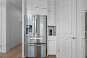 Kitchen with light wood finished floors, tasteful backsplash, stainless steel refrigerator with ice dispenser, baseboards, and white cabinets