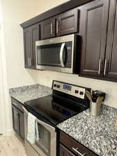 Kitchen with dark brown cabinetry, light stone countertops, light wood-style floors, and appliances with stainless steel finishes