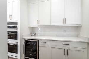 Bar with white cabinets, beverage cooler, light stone counters, double oven, and decorative backsplash