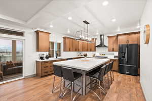 Kitchen with a large island, black fridge with ice dispenser, wall chimney range hood, and light wood finished floors