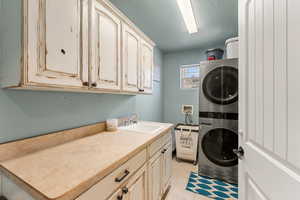 Laundry area featuring a textured ceiling, a sink, cabinet space, stacked washing maching and dryer, and light tile patterned floors