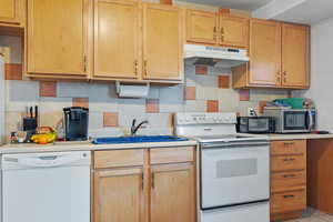 Kitchen featuring light countertops, white appliances, under cabinet range hood, decorative backsplash, and a sink