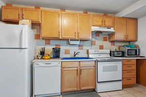 Kitchen featuring light countertops, white appliances, under cabinet range hood, decorative backsplash, and a sink