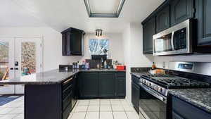 Kitchen featuring light tile patterned floors, stainless steel appliances, dark stone counters, a sink, and a peninsula