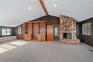 Unfurnished living room featuring wood walls, carpet flooring, lofted ceiling with beams, visible vents, and a stone fireplace