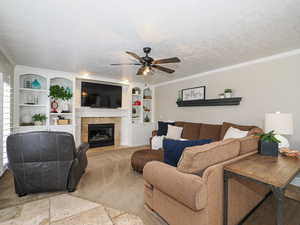 Carpeted living area with crown molding, a tiled fireplace, a ceiling fan, a textured ceiling, and built in features