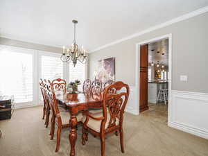 Dining area with a wainscoted wall, light colored carpet, ornamental molding, and an inviting chandelier