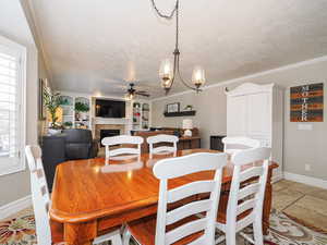 Dining room with crown molding, ceiling fan with notable chandelier, a textured ceiling, a tiled fireplace, and stone tile flooring