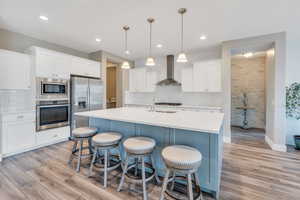 Kitchen with light countertops, a breakfast bar area, wall chimney exhaust hood, light wood-type flooring, and stainless steel appliances