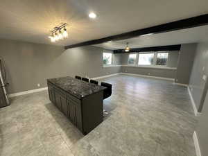 Kitchen featuring ceiling fan, a kitchen island, dark stone countertops, baseboards, and beam ceiling
