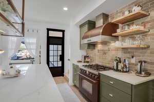 Kitchen with high end stainless steel range, backsplash, open shelves, green cabinets, and wall chimney exhaust hood