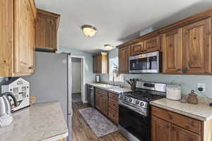 Kitchen featuring stainless steel appliances, a sink, brown cabinetry, light countertops, and dark wood-type flooring