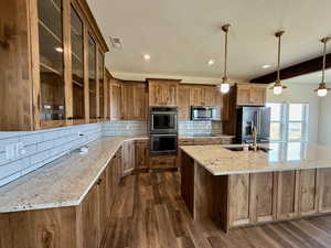 Kitchen with decorative backsplash, glass insert cabinets, brown cabinets, light stone countertops, and decorative light fixtures