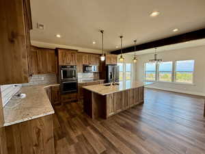 Kitchen with decorative backsplash, light stone countertops, a kitchen island with sink, pendant lighting, and beam ceiling