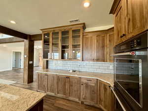 Kitchen featuring glass insert cabinets, decorative backsplash, recessed lighting, light stone countertops, and dark wood-type flooring