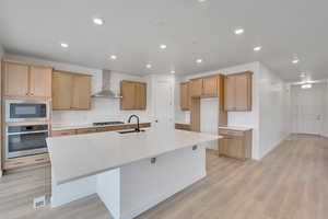 Kitchen featuring light wood-style floors, an island with sink, wall chimney range hood, a sink, and stainless steel appliances