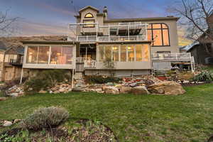 Back of house at dusk featuring a balcony, a wooden deck, a sunroom, and a lawn