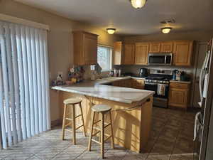 Kitchen with stainless steel appliances, a peninsula, light countertops, a breakfast bar, and brown cabinetry