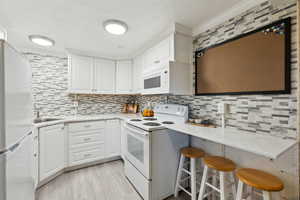Mother-in-law Kitchen with light wood-style floors, white appliances, tasteful backsplash, white cabinets, and a breakfast bar area