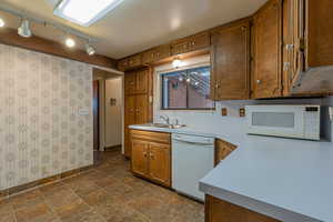 Kitchen featuring wallpapered walls, white appliances, stone finish floor, brown cabinetry, and a sink