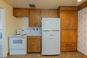 Kitchen featuring wallpapered walls, white appliances, light countertops, brown cabinetry, and stone finish flooring
