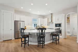 Kitchen with wall chimney range hood, appliances with stainless steel finishes, light wood-style floors, white cabinetry, and a kitchen island
