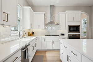 Kitchen with wall chimney exhaust hood, white cabinets, light countertops, a sink, and stainless steel appliances