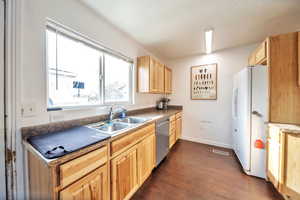 Kitchen with freestanding refrigerator, light brown cabinetry, dishwasher, dark countertops, and a sink