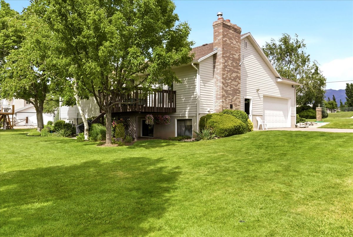 Expansive lawn with mature trees, a multi-level deck, white vinyl siding, a prominent brick chimney, and an attached two-car garage