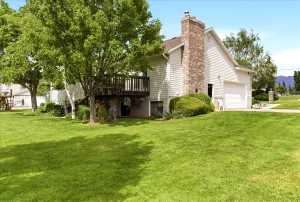 Expansive lawn with mature trees, a multi-level deck, white vinyl siding, a prominent brick chimney, and an attached two-car garage