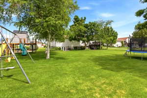 Spacious green lawn with mature trees, a multi-level deck, and a play structure featuring a slide
