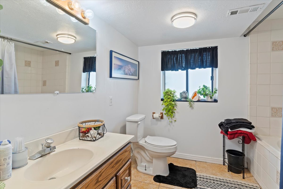 Bathroom featuring a single-sink vanity with light-colored countertop and wood-finish cabinetry, a window with black privacy curtain, a toilet, and a tiled shower/tub combination