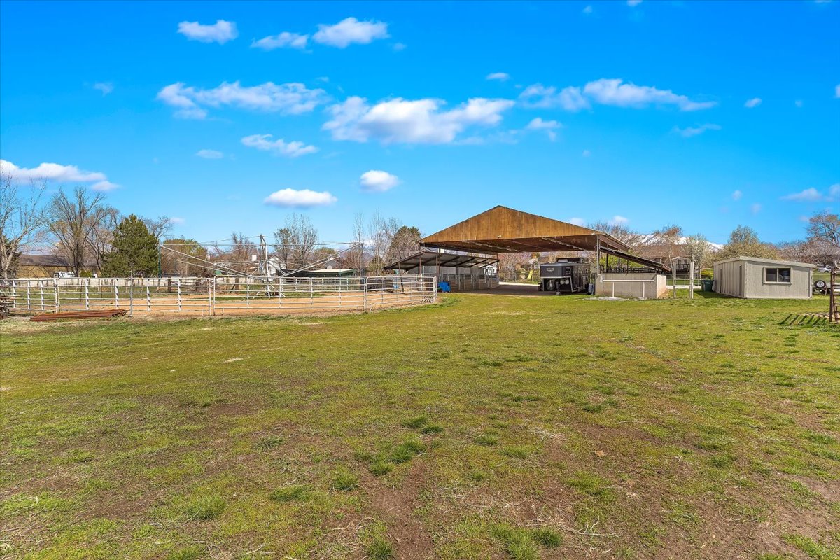Expansive green space featuring a covered outdoor structure, a fenced arena, a detached shed, and a mountain backdrop