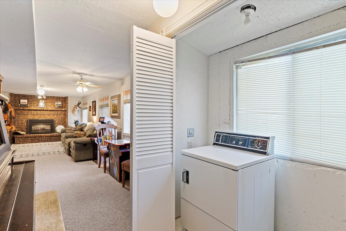 Dedicated laundry area with a large window and louvered bifold door, offering direct access to a carpeted living space featuring a brick fireplace with an insert and ceiling fans