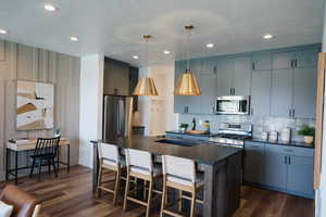 Kitchen featuring dark countertops, appliances with stainless steel finishes, a kitchen island with sink, and dark wood-style flooring