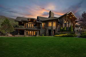 Back of property at dusk featuring stone siding, a chimney, a lawn, a balcony, and stairs