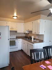 Kitchen featuring white appliances, white cabinets, under cabinet range hood, a sink, and dark countertops
