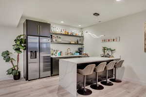 Bar with indoor wet bar, baseboards, light wood-style flooring, stainless steel refrigerator with ice dispenser, and a sink