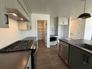 Kitchen with dark wood-style flooring, dark countertops, stainless steel appliances, decorative light fixtures, and backsplash