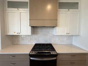 Kitchen featuring stainless steel gas stove, wall chimney range hood, tasteful backsplash, glass insert cabinets, and light stone counters