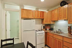 Apartment kitchen featuring baseboard heating, white appliances, backsplash, brown cabinets, and a sink
