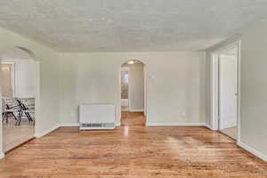 Empty room featuring heating unit, a textured ceiling, arched walkways, light wood-style flooring, and baseboards