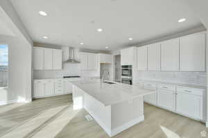 Kitchen featuring wall chimney range hood, recessed lighting, backsplash, light wood finished floors, and double oven